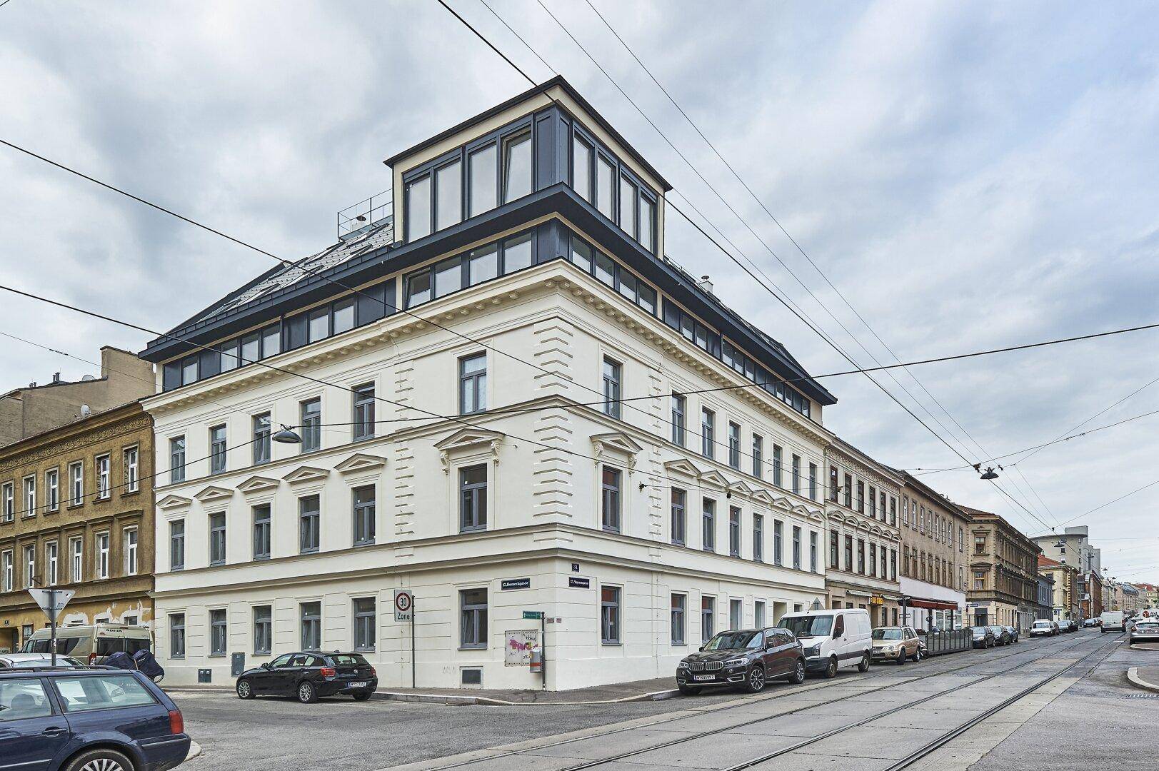 Eckgebäude mit historischer Fassade und moderner Dachaufstockung, an einer belebten Stadtkreuzung gelegen.