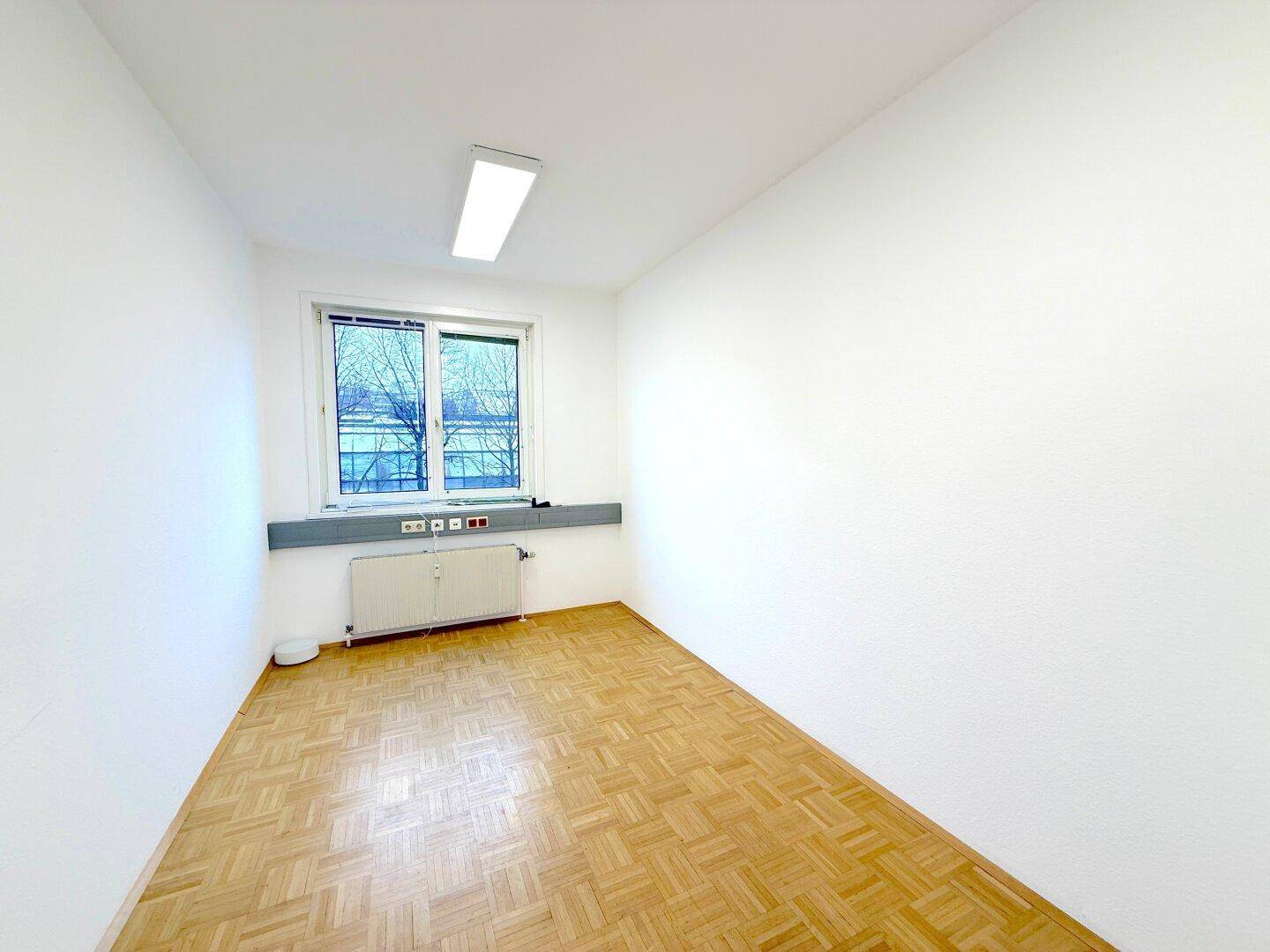 Leerer Büroraum mit hellem Parkettboden, weißen Wänden und einem Fenster mit Heizung darunter.