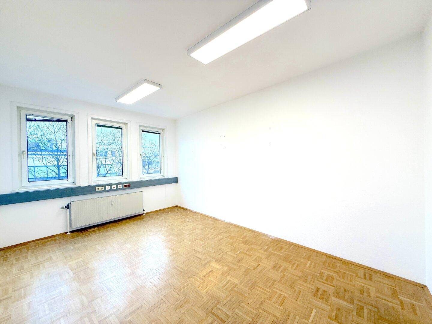 Geräumiger, leerer Büroraum mit drei Fenstern, weißen Wänden und Fischgrätparkett.