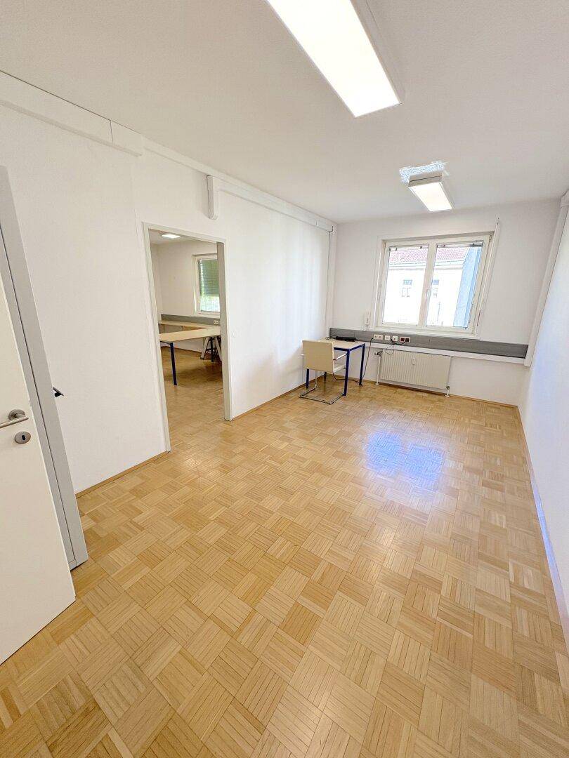 Leerer Büroraum mit Parkettboden, einem Fenster und einer Verbindungstür zu einem weiteren Raum.