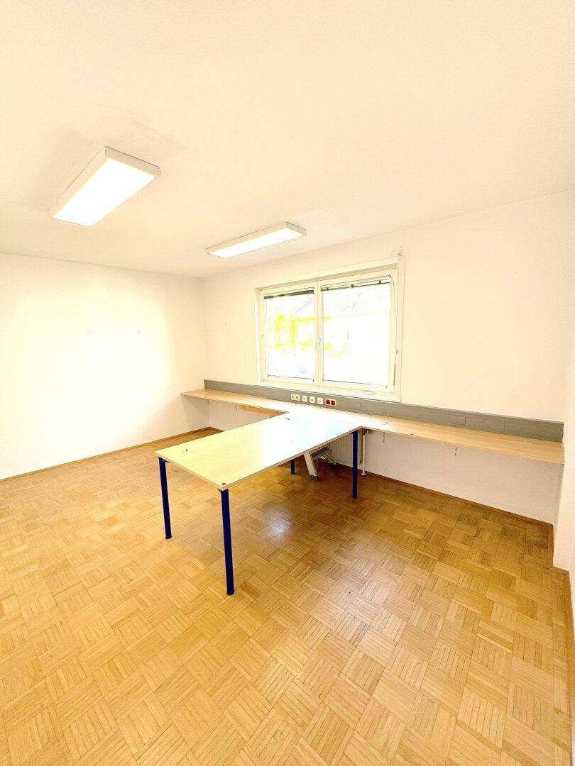 Einfacher Büroraum mit einem Tisch, einer langen Ablagefläche unter dem Fenster und Parkettboden.
