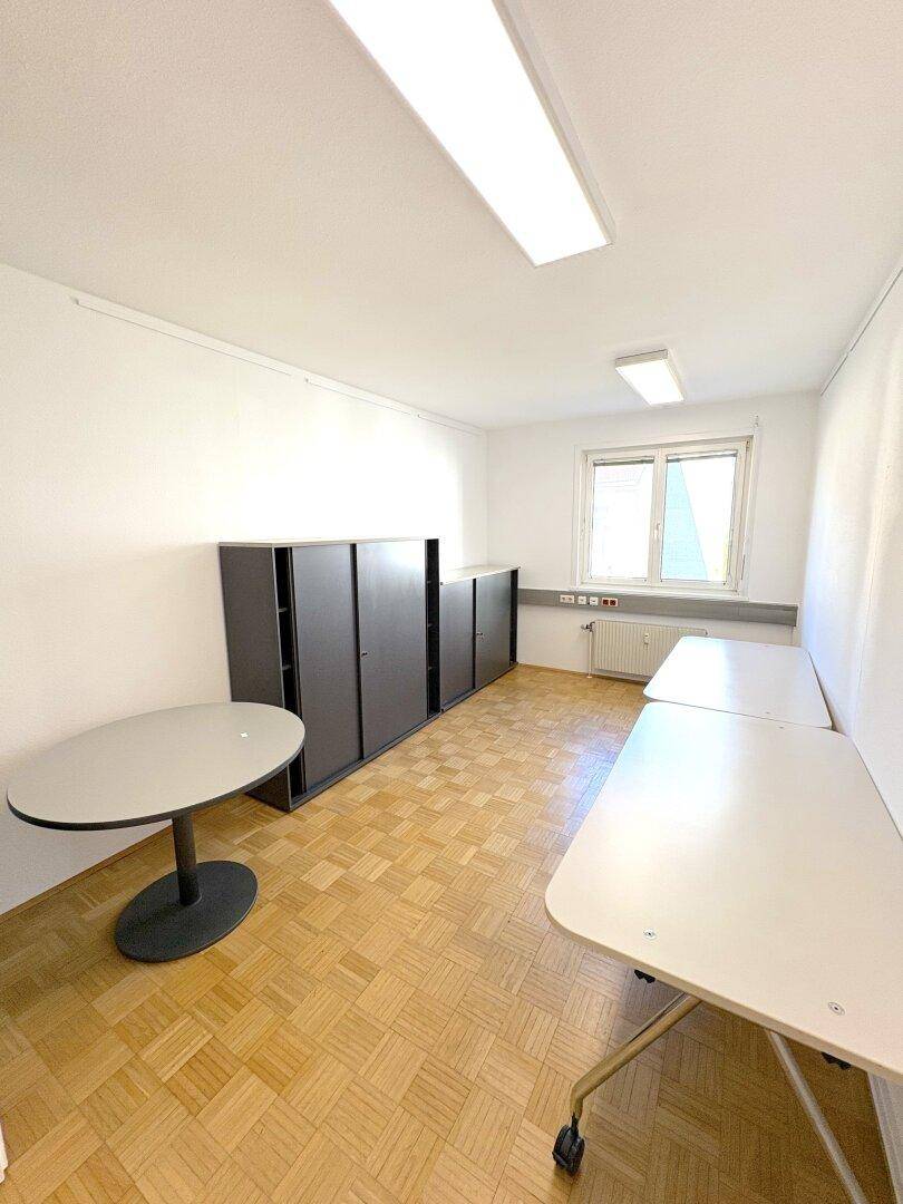 Büroraum mit mehreren Tischen, grauen Schränken und Parkettboden.