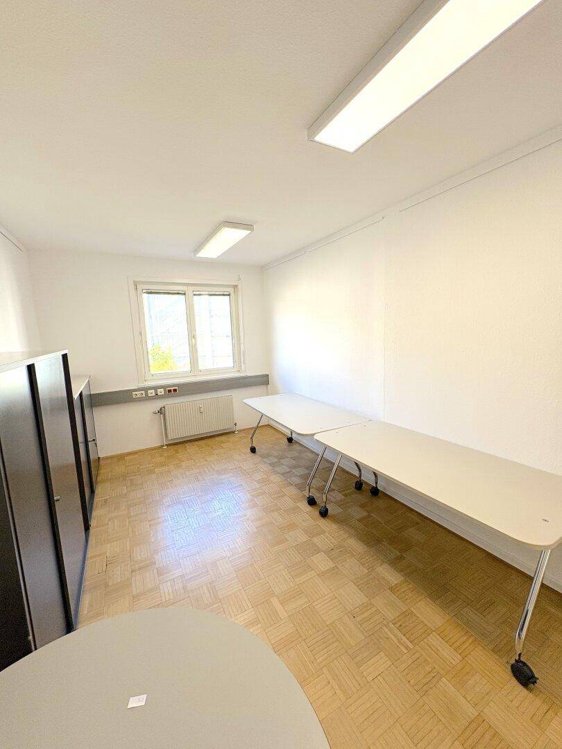 Büroraum mit langen Tischen auf Rollen, grauen Schränken und einem Fenster.