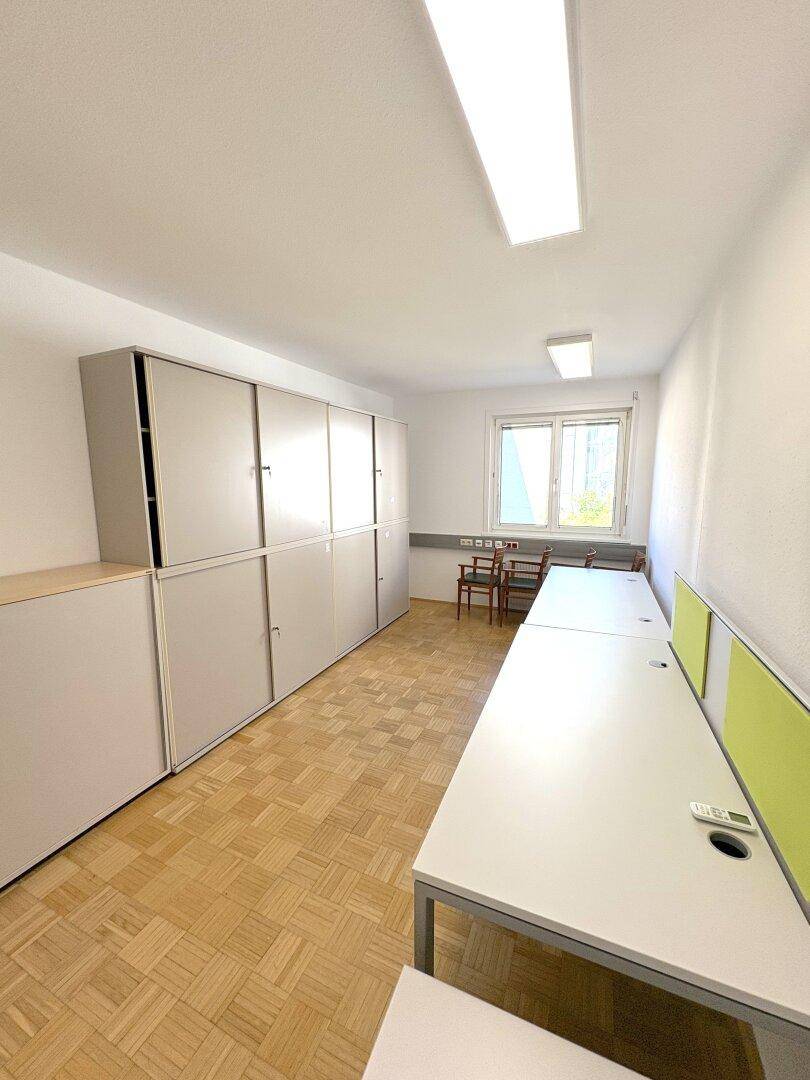 Büroraum mit langen Tischen, hellgrauen Schränken und einem Fenster.