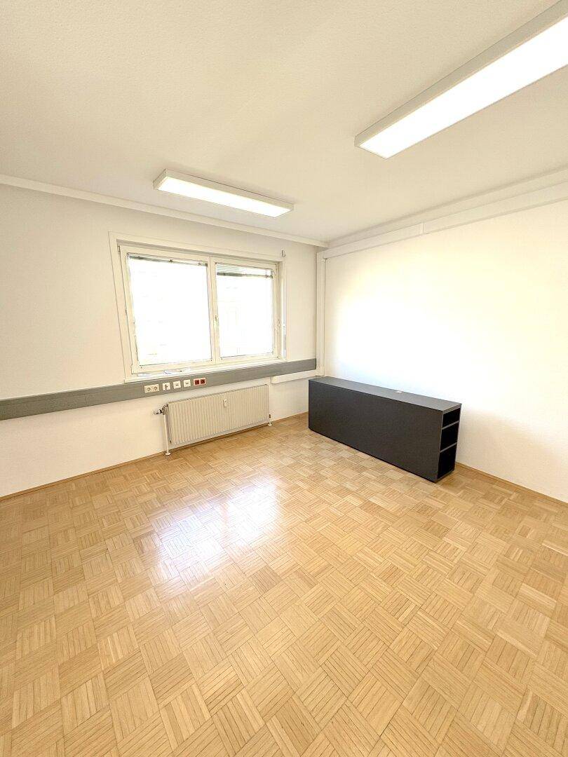 Leerer Büroraum mit Parkettboden, einem Fenster und einem dunklen Regal.