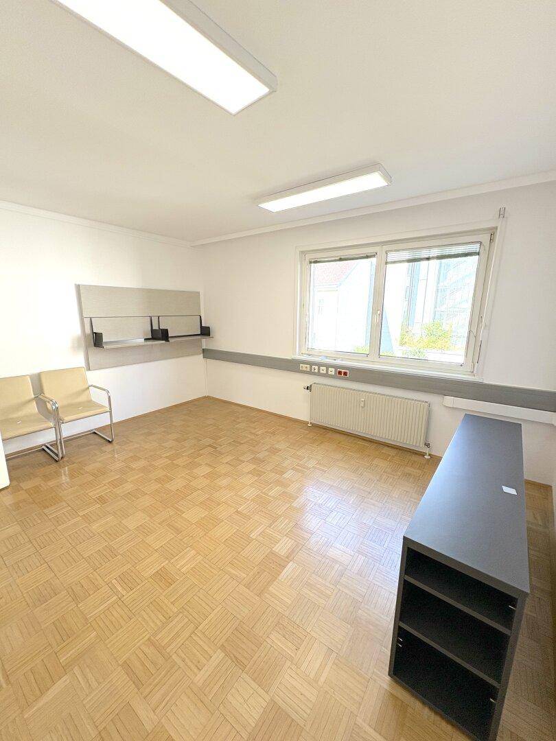 Leerer Büroraum mit Parkettboden, einem Fenster und einem dunklen Regal.