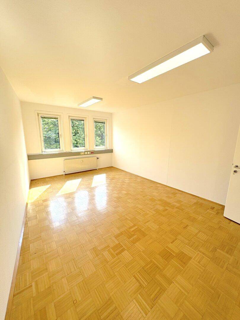 Leerer Büroraum mit Parkettboden, drei Fenstern und viel natürlichem Licht.
