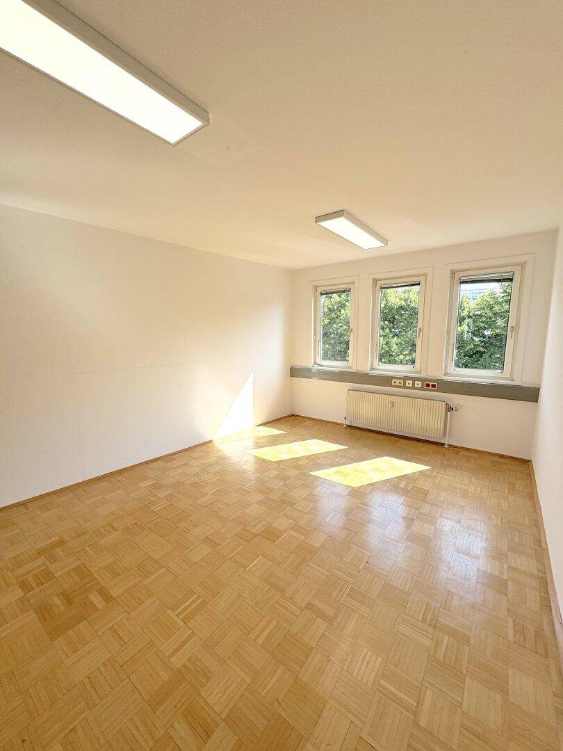 Leerer Büroraum mit Parkettboden, drei Fenstern und viel natürlichem Licht.