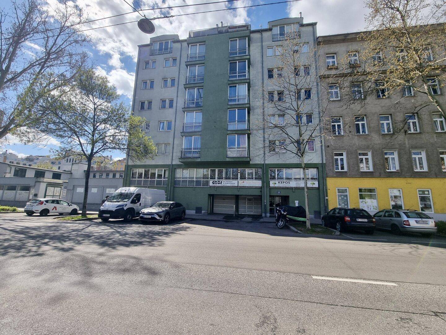 Ansicht des Wohngebäudes von der Straße, umgeben von Bäumen und anderen Gebäuden.