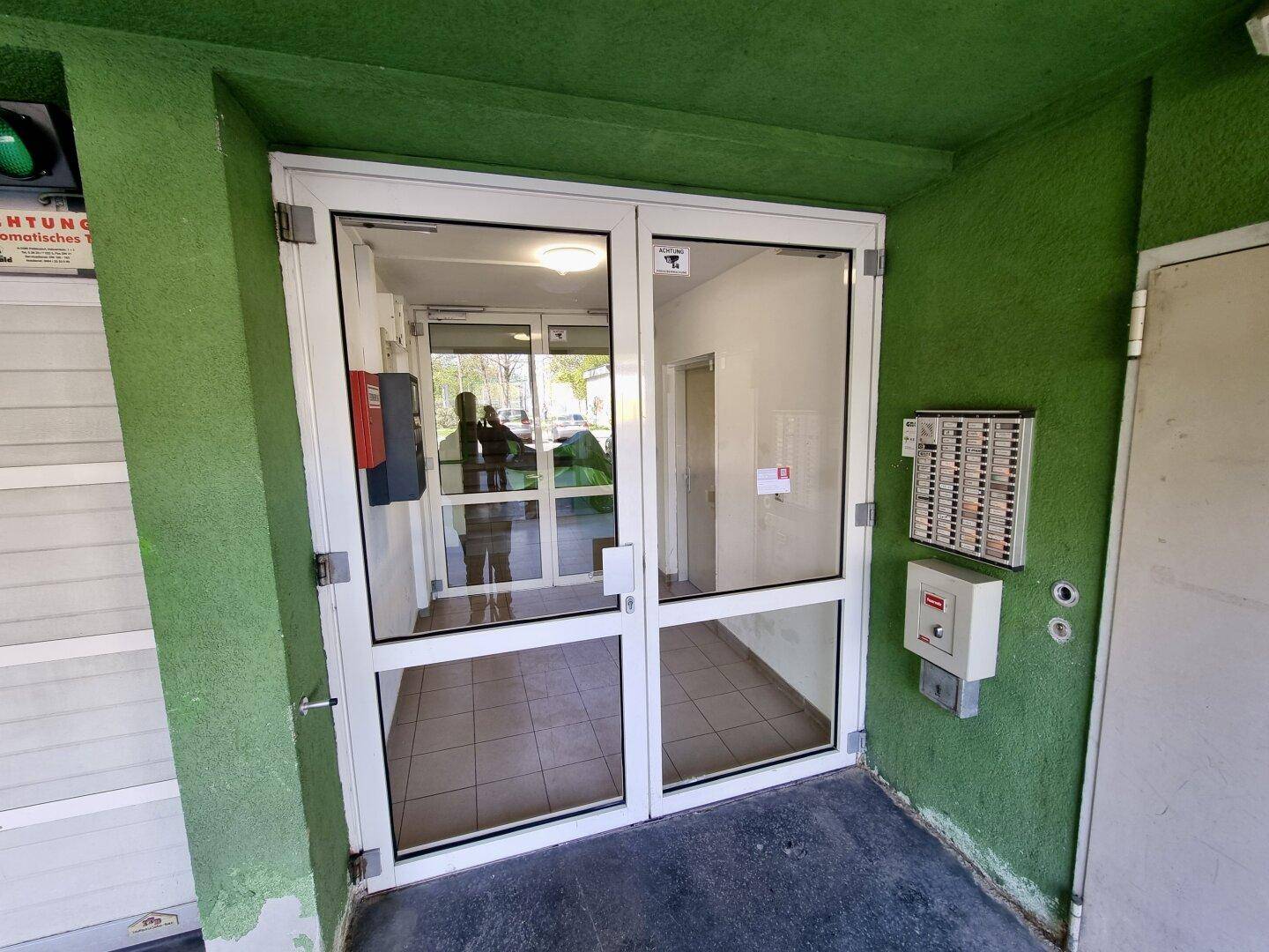 Haupteingang des Gebäudes mit Glastüren und Gegensprechanlage, in grüner Fassade.