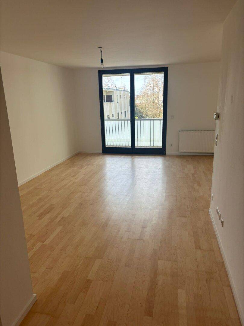 Geräumiges Wohnzimmer mit hellem Parkettboden und großen Fenstern, die viel Tageslicht hereinlassen.