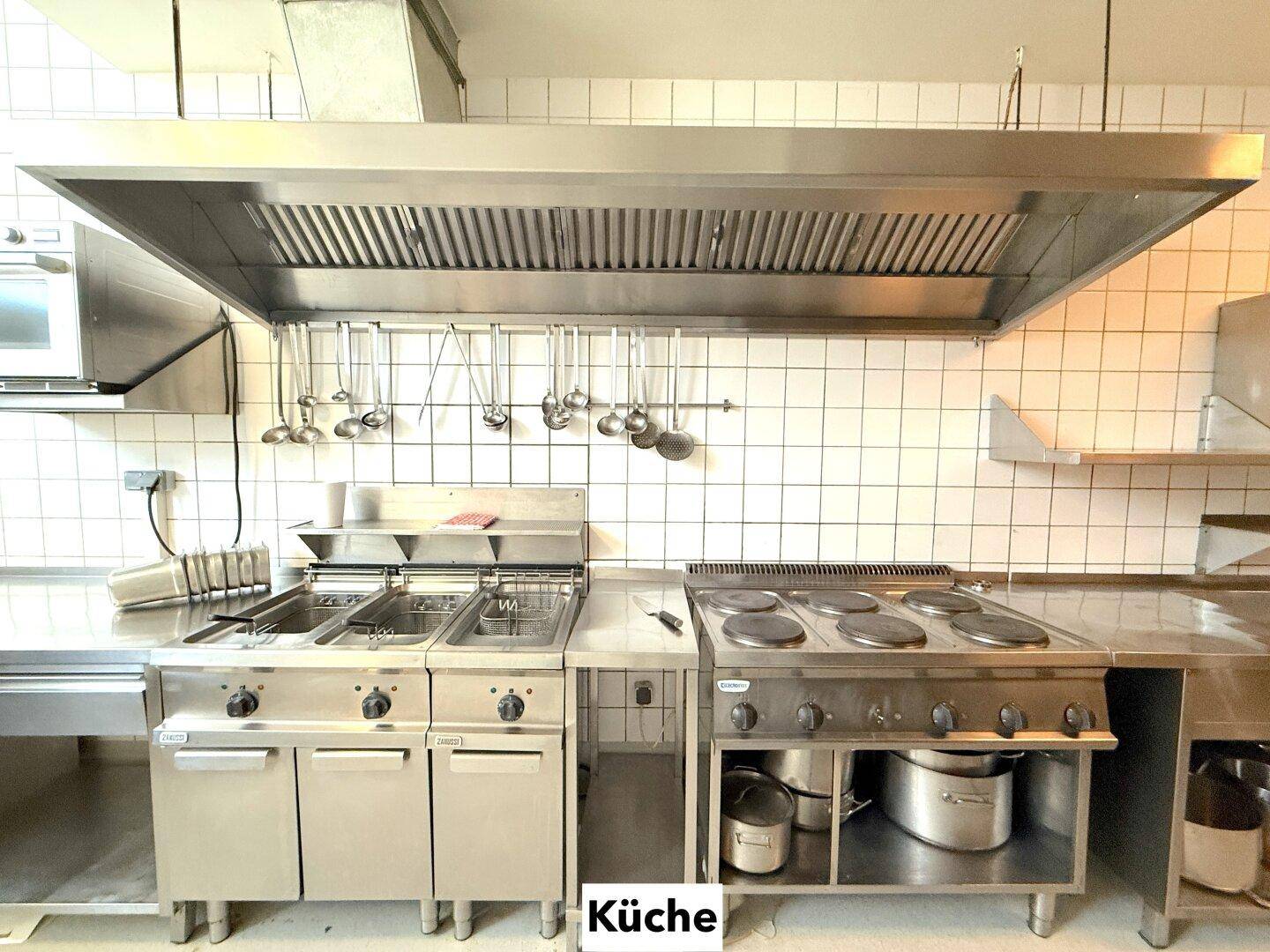 Professionelle Großküche mit Edelstahlgeräten, Fritteusen, Kochfeldern und einer großen Dunstabzugshaube.