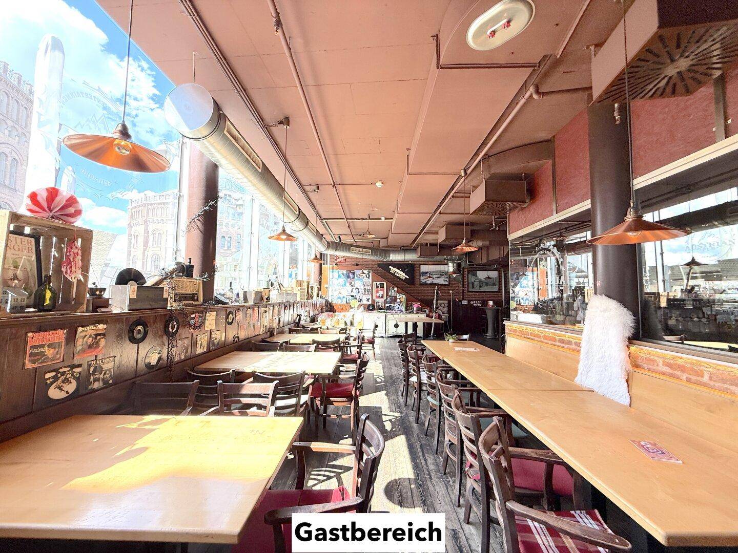 Langer Gastbereich mit Holztischen, Bänken und Stühlen, beleuchtet durch große Fenster und Hängelampen.