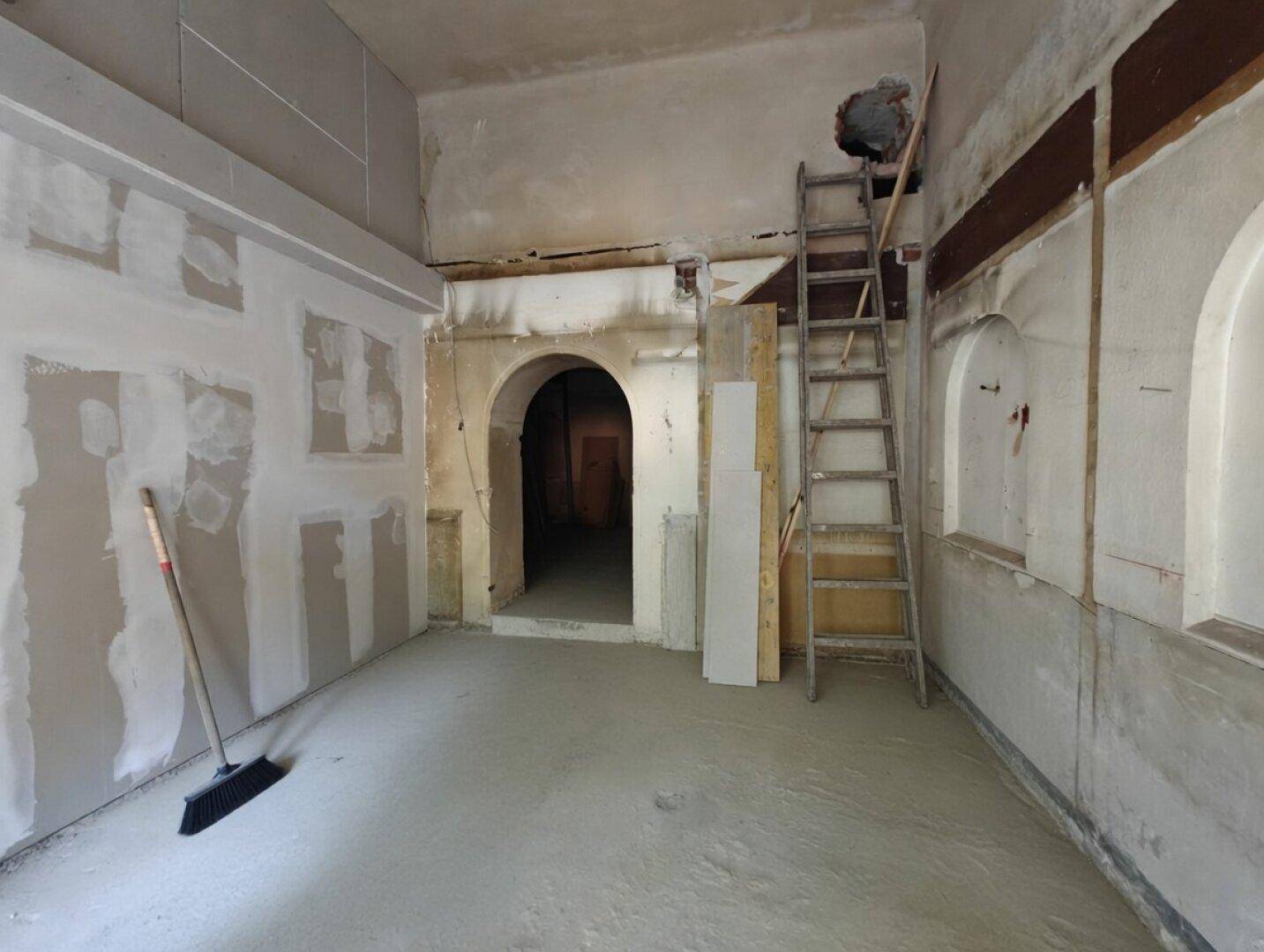 Innenraum im Rohbau mit Trockenbauwänden, Betonboden und einem Bogen als Durchgang.