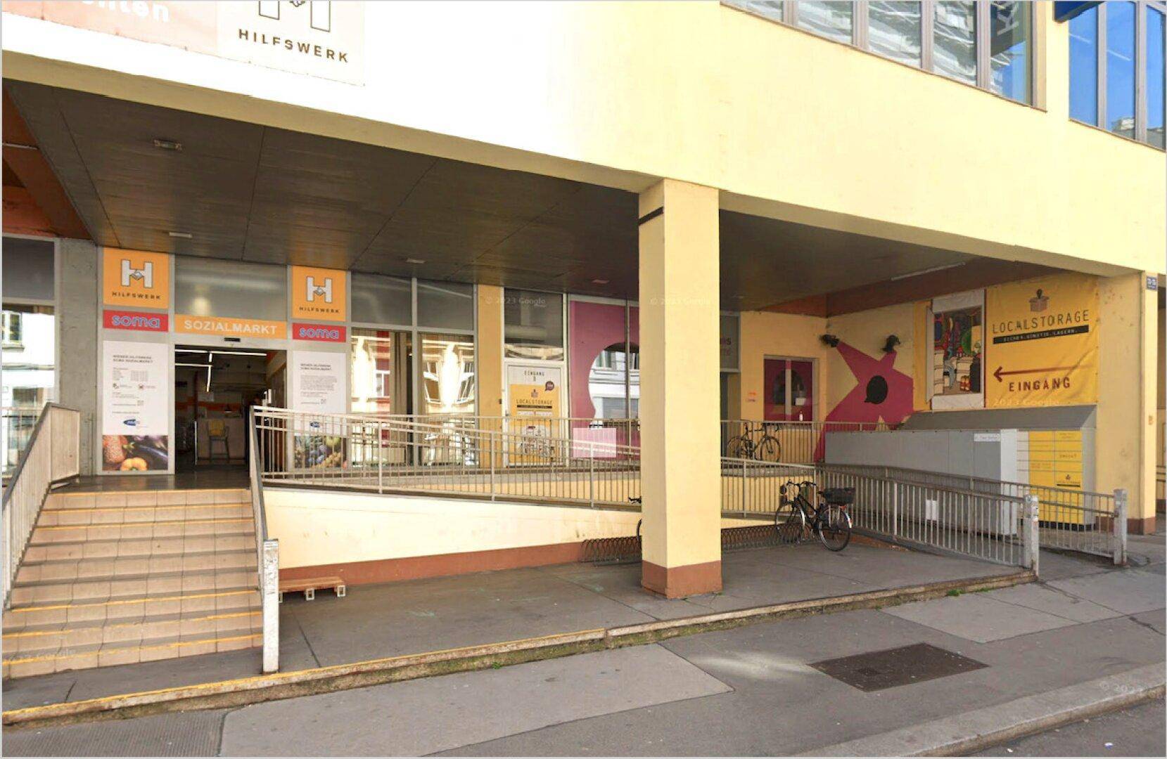 Gebäudeeingang mit Rampe und Treppe, der zu einem Sozialmarkt und Lagerflächen führt.