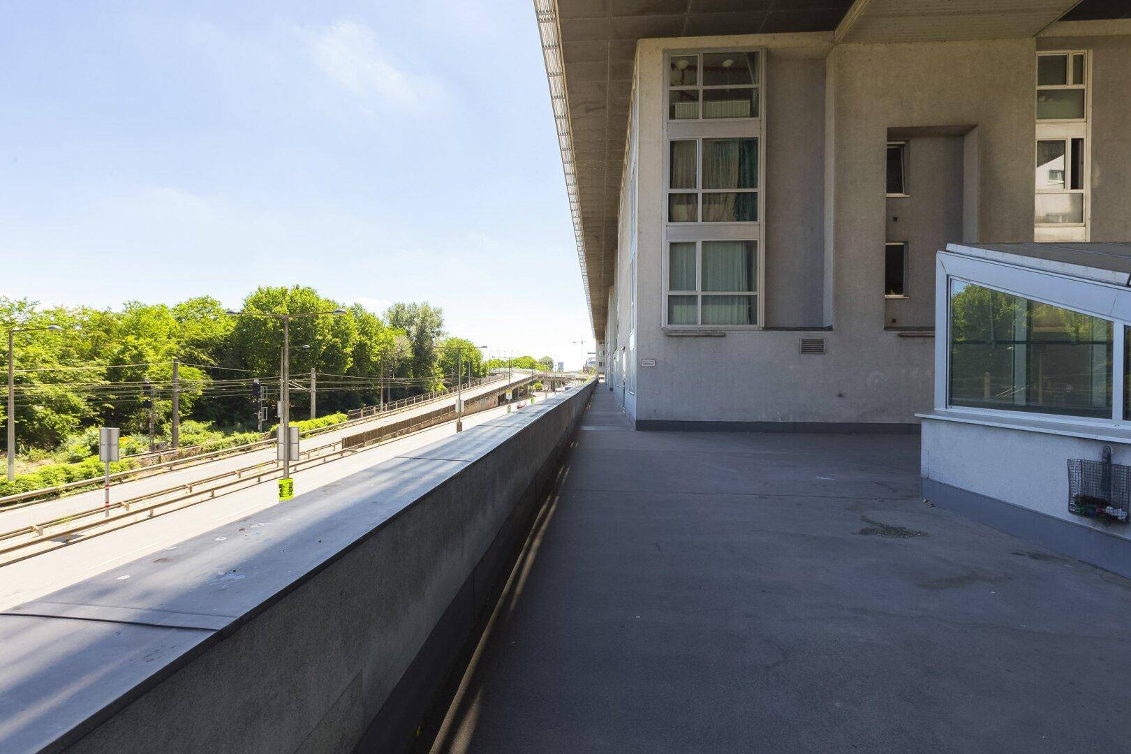 Langer Gehweg entlang eines Gebäudes mit Blick auf eine Straße und Bäume.