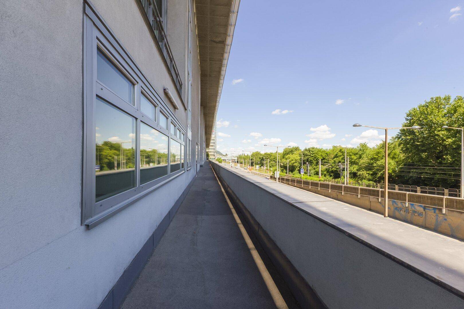 Langer Gehweg entlang eines Gebäudes mit Blick auf eine Straße und Bäume.