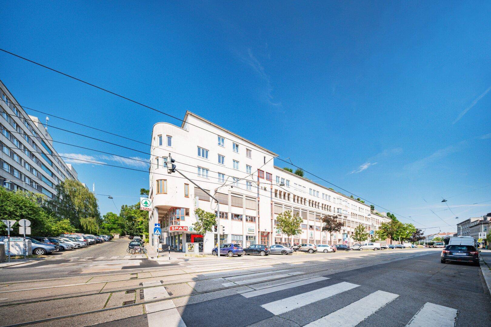 Modernes Gebäude mit weißer Fassade und großen Fenstern an einer belebten Stadtstraße unter blauem Himmel.