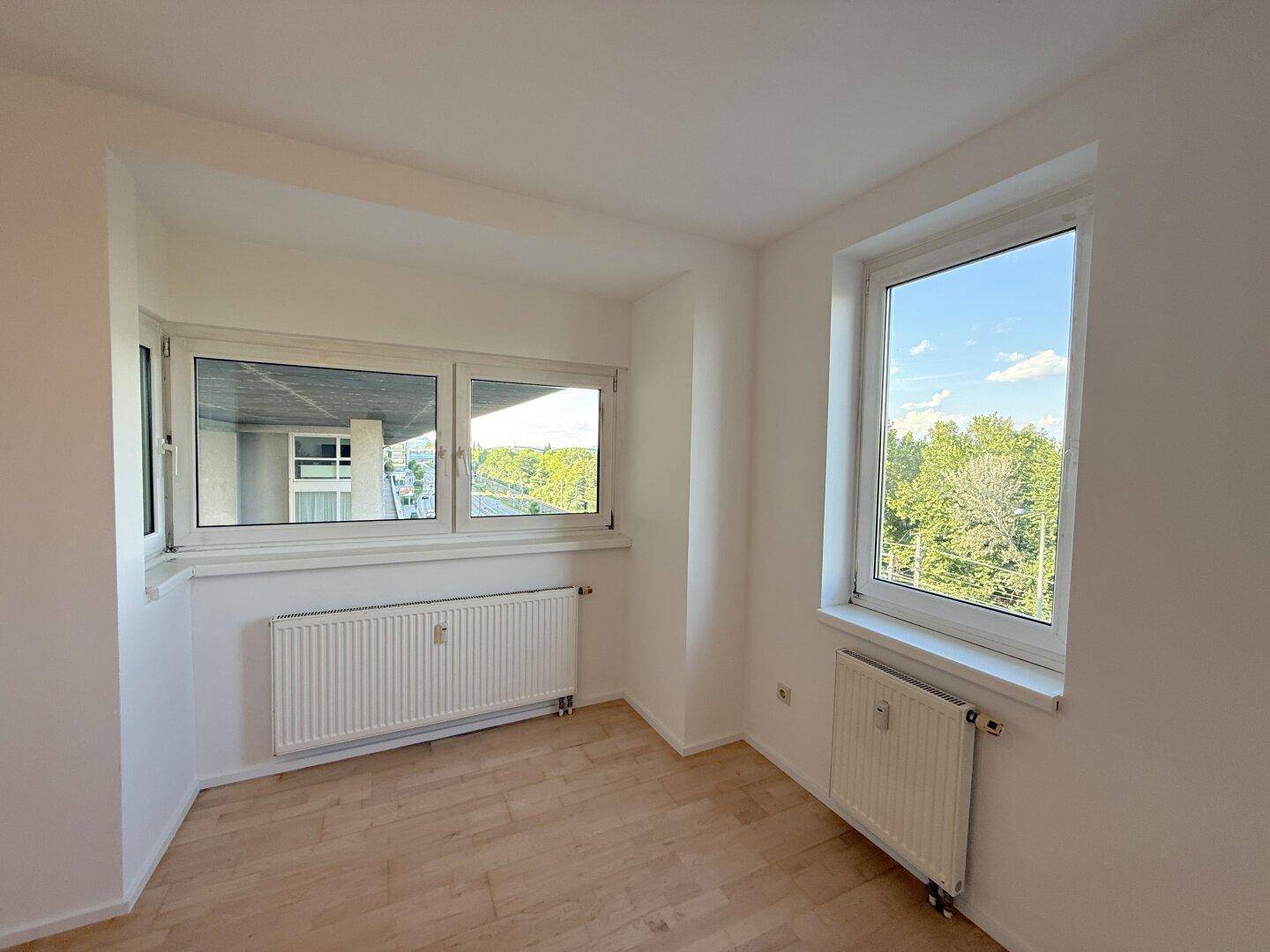 Helles Schlafzimmer mit Holzfußboden und zwei Fenstern, die einen Ausblick ins Freie bieten.