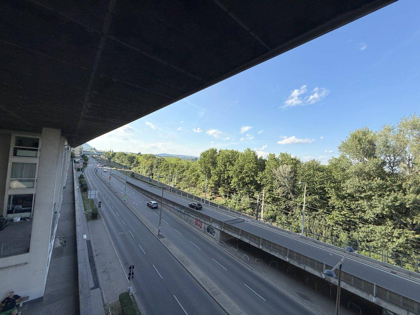 Weitläufiger Blick auf eine Straße und grüne Bäume von einem erhöhten Standpunkt aus.