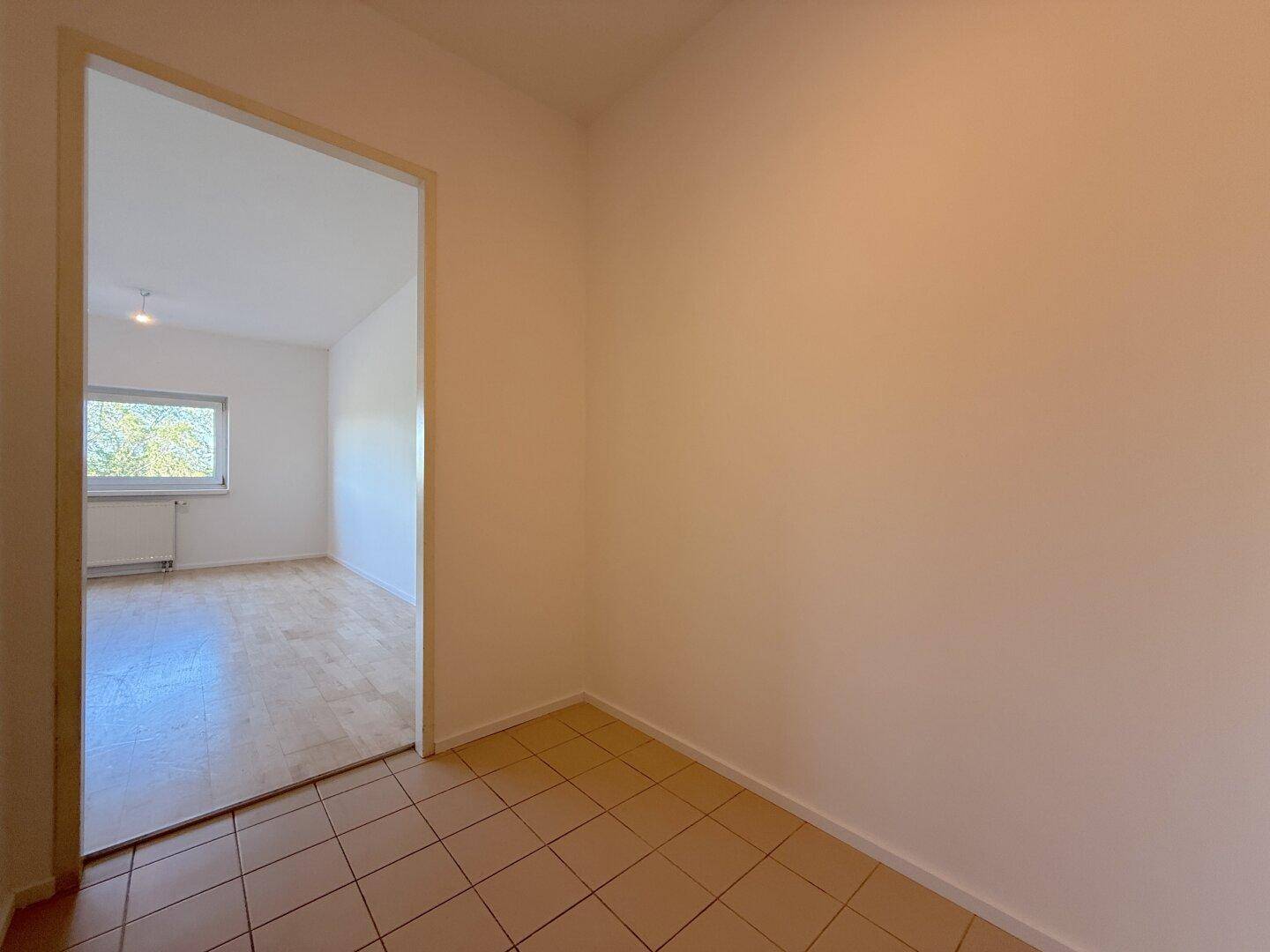 Flur mit braunen Fliesen, der zu einem weiteren hellen Zimmer mit Holzfußboden führt.