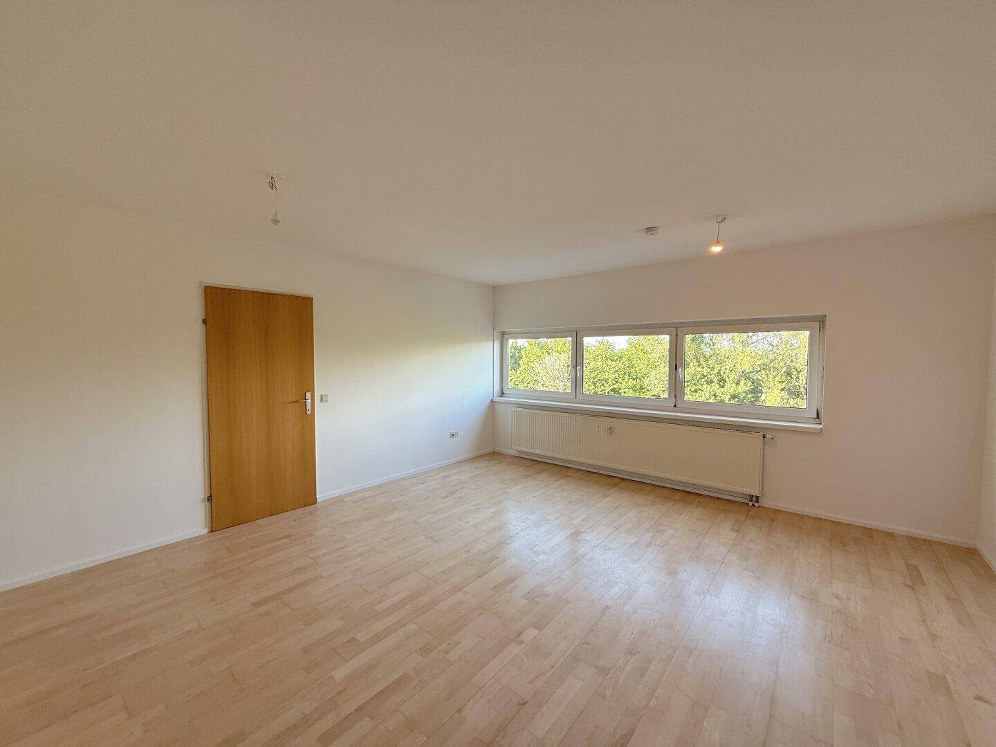 Geräumiges Wohnzimmer mit hellem Holzfußboden, einer Holztür und Fenstern mit Grünblick.