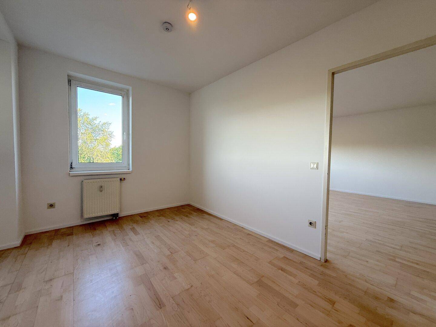Leeres Zimmer mit Holzfußboden und einem Fenster, das viel natürliches Licht hereinlässt.