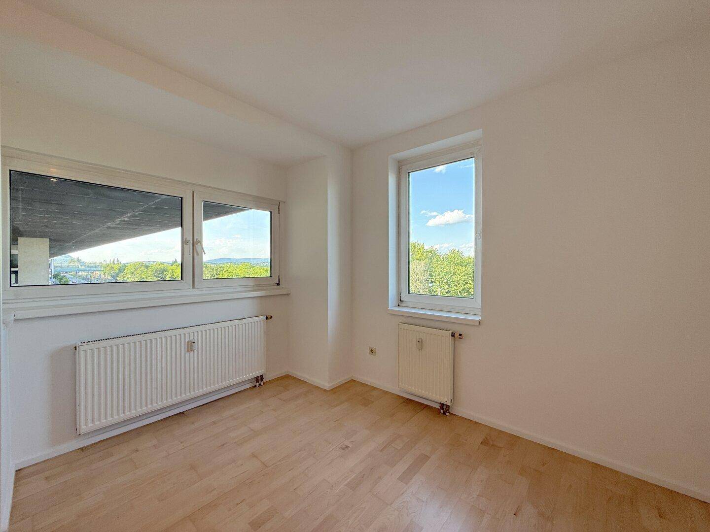 Helles Schlafzimmer mit Holzfußboden und zwei Fenstern, die einen Ausblick ins Freie bieten.