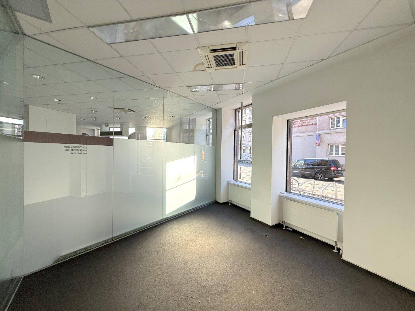 Kleinerer Bürobereich mit Glaswand, Blick auf die Straße und dunklem Bodenbelag.