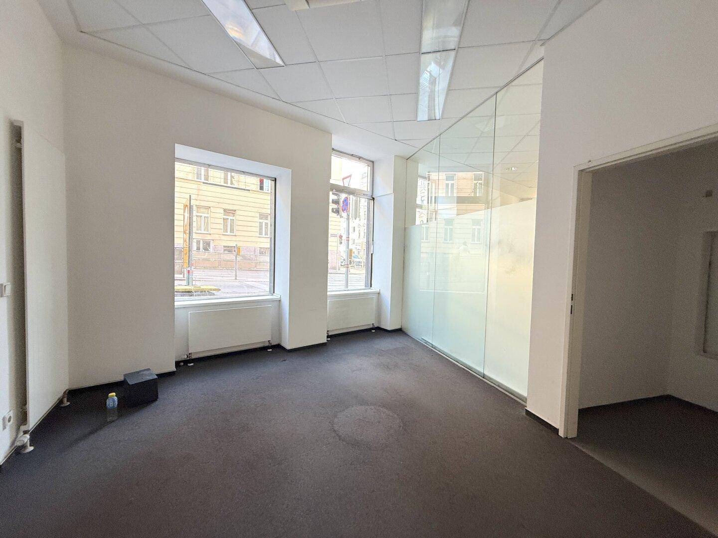 Heller Büroraum mit zwei großen Fenstern, dunklem Teppichboden und einer Glasabtrennung.
