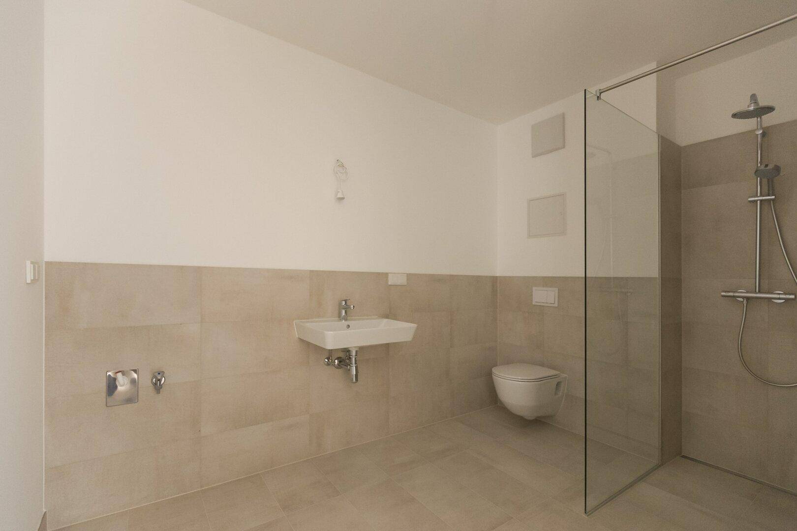 Modernes Badezimmer mit beigefarbenen Fliesen, wandhängendem WC, Waschbecken und einer geräumigen begehbaren Dusche.