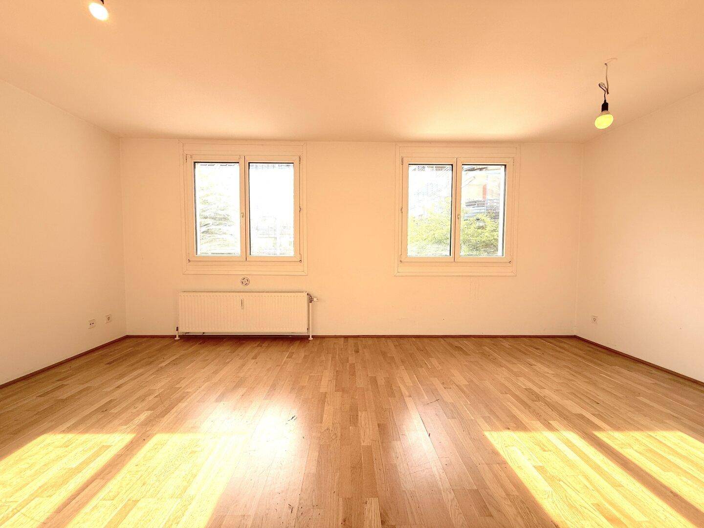 Geräumiges Zimmer mit hellem Parkettboden und zwei großen Fenstern, die den Raum erhellen.