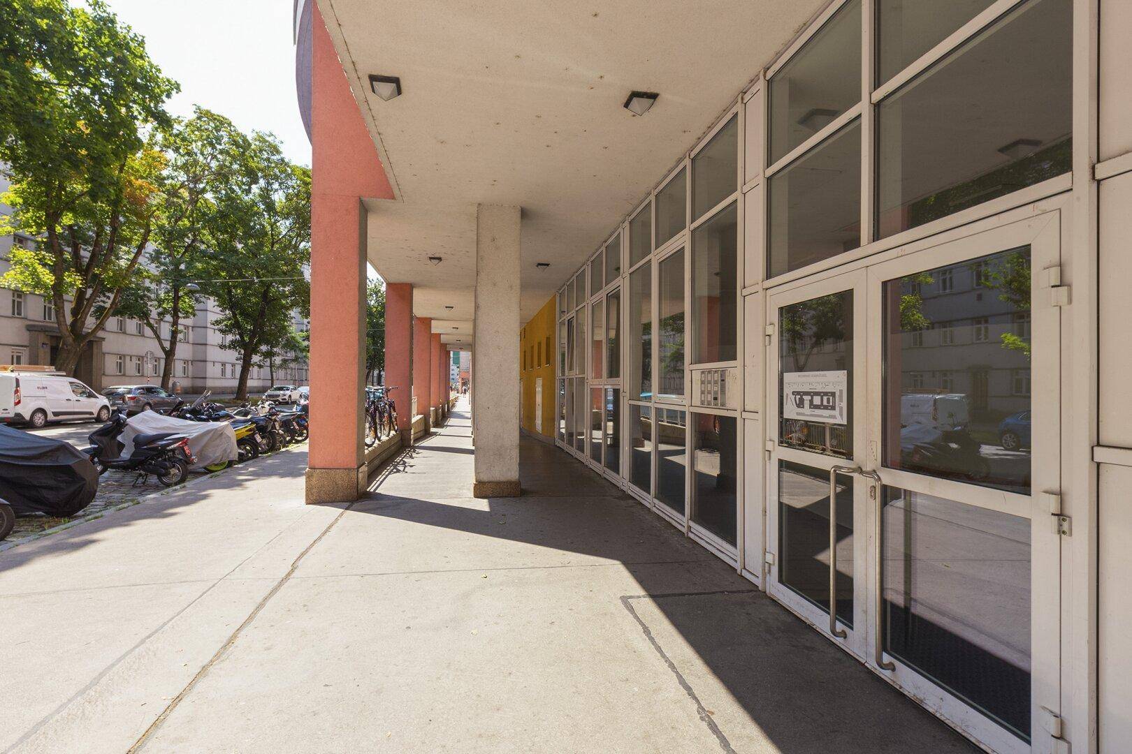 Überdachter Gehweg entlang einer Gebäudefassade mit großen Fenstern und Geschäften.