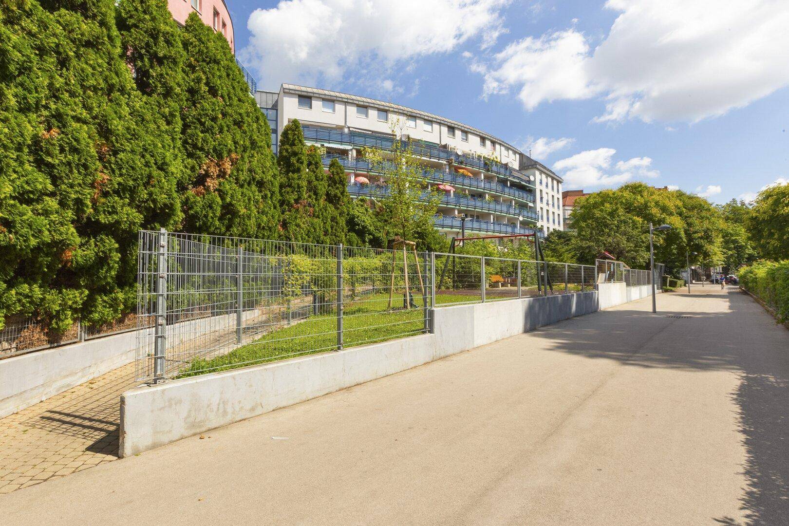 Außenbereich mit Gehweg, Zaun und Grünflächen, umgeben von Wohngebäuden.