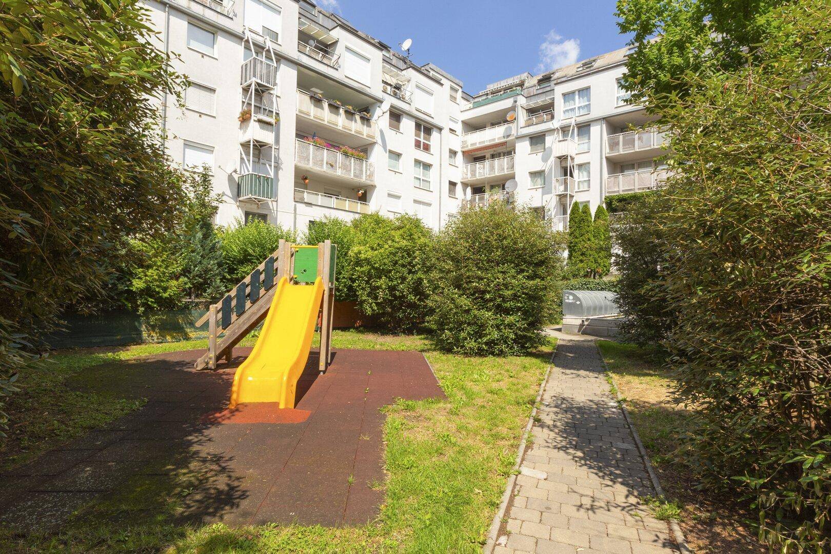 Spielplatz mit gelber Rutsche und Grünflächen im Innenhof einer modernen Wohnanlage.