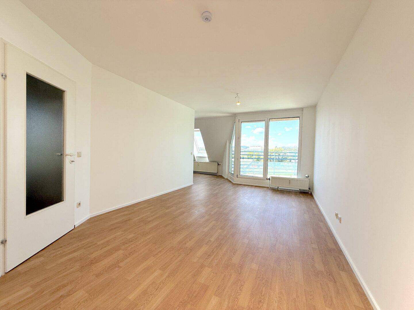 Helles, leeres Wohnzimmer mit Laminatboden und großen Fenstern mit Weitblick.