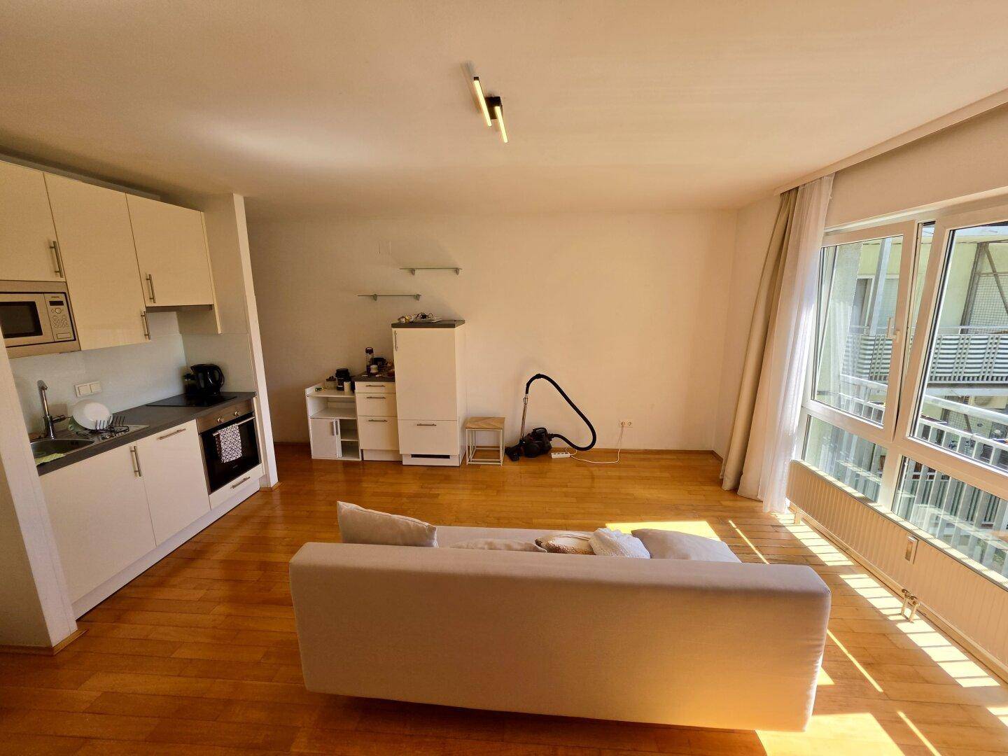 Heller Wohnbereich mit Küchenzeile, Sofa und großem Fenster, das viel Licht hereinlässt.