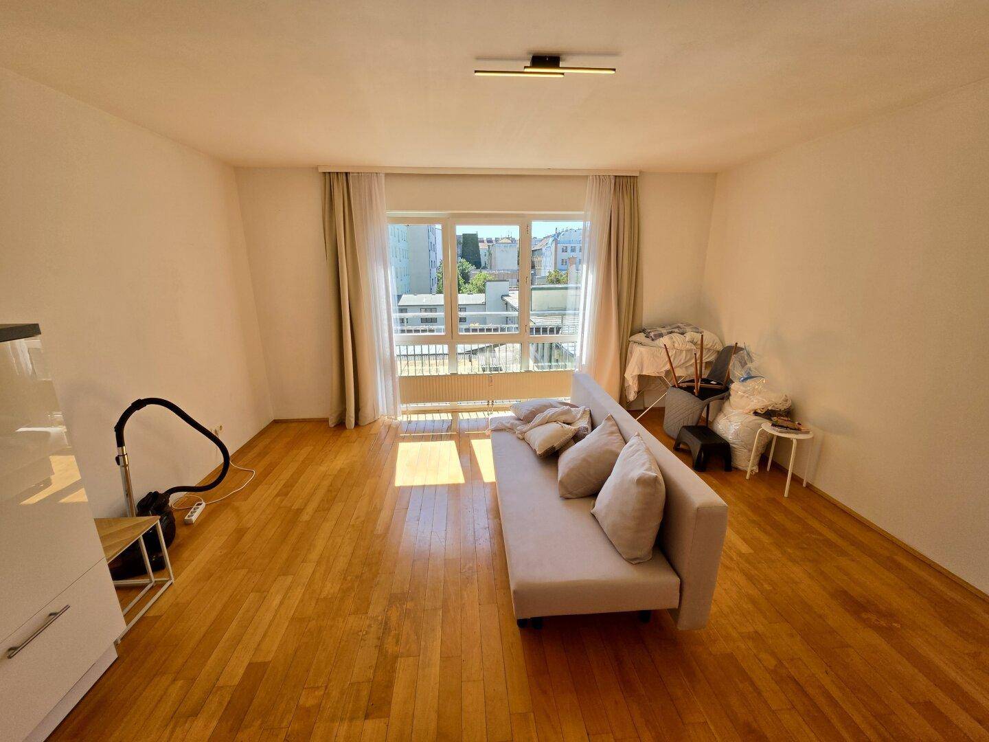 Helles Wohnzimmer mit Parkettboden, beigem Sofa und großem Fenster mit Blick nach draußen.