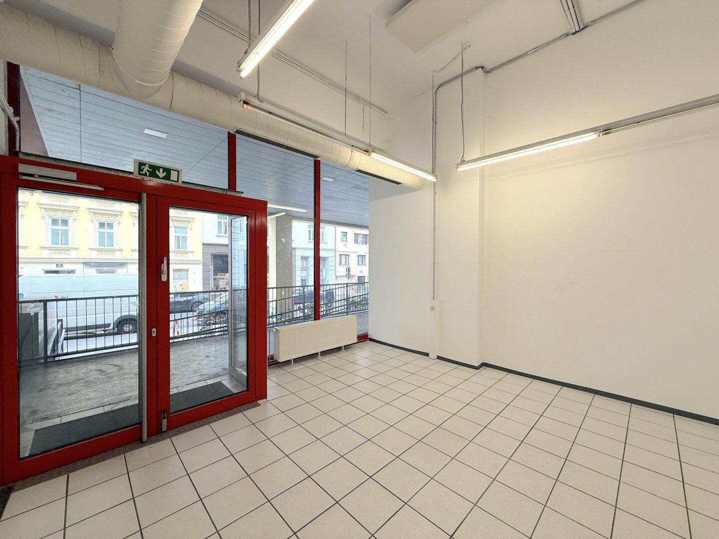 Heller Gewerberaum mit roten Eingangstüren, Fliesenboden und viel Tageslicht von der Fensterfront.