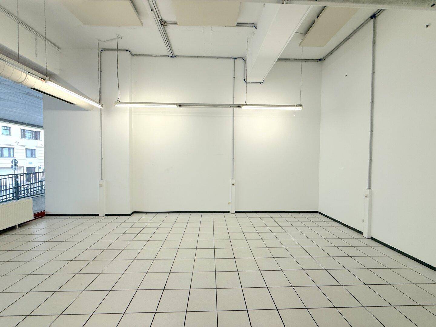 Geräumiger, leerer Gewerberaum mit weißen Wänden, Fliesenboden und sichtbaren Deckeninstallationen.