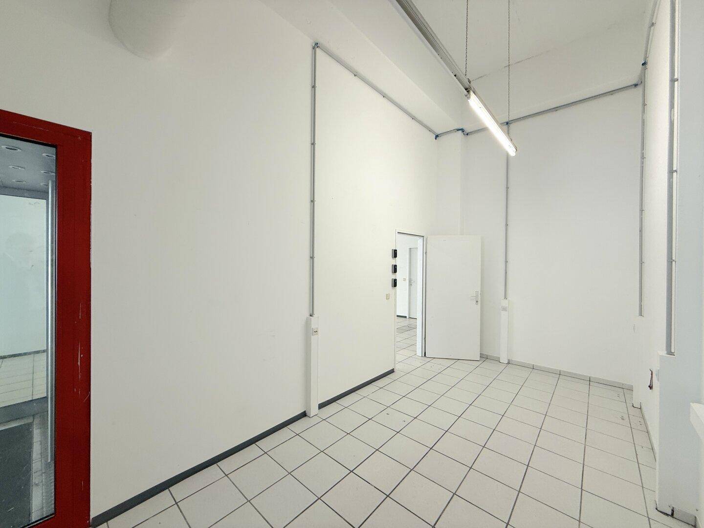 Heller Gewerberaum mit weiß gestrichenen Wänden, Fliesenboden und einer Tür zu einem Nebenraum.