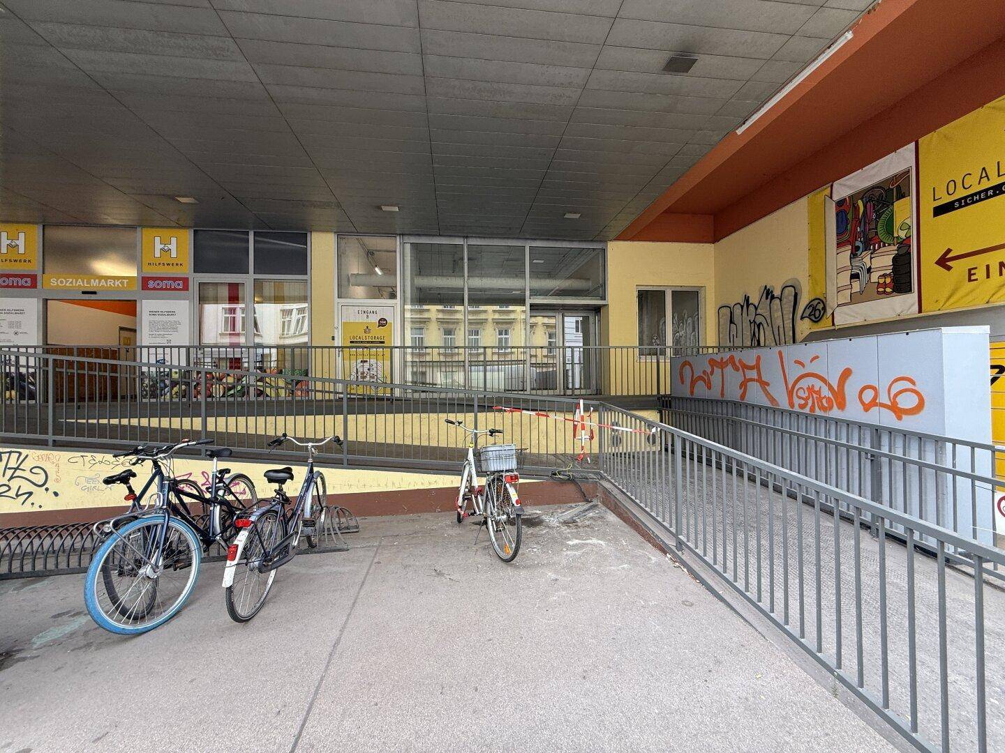 Außenbereich des Gebäudes mit barrierefreier Rampe, Fahrradstellplätzen und Blick auf die Ladenlokale.