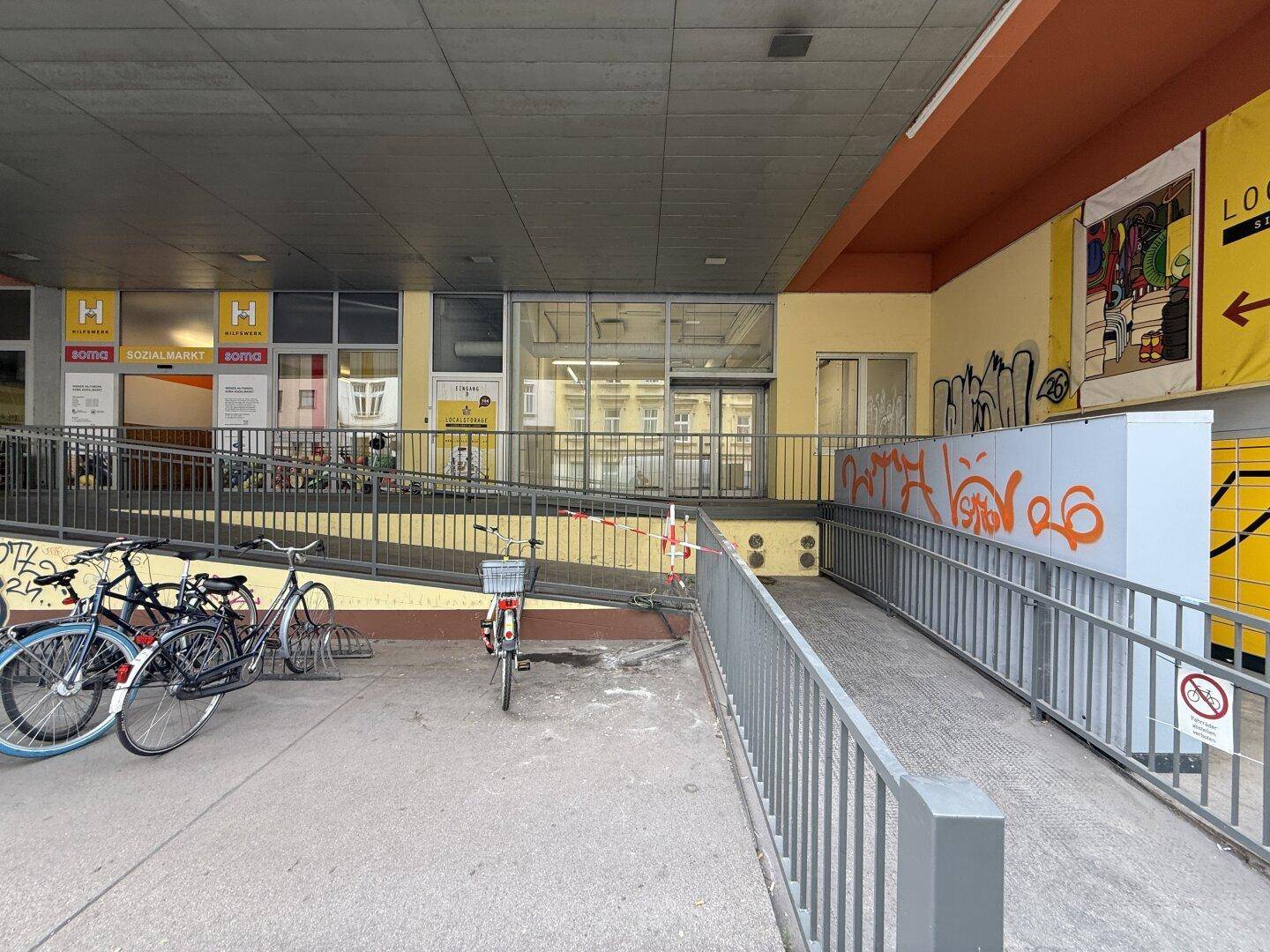 Gebäudeeingang mit Rampe, Fahrradabstellmöglichkeiten und Blick auf die umliegenden Geschäfte.