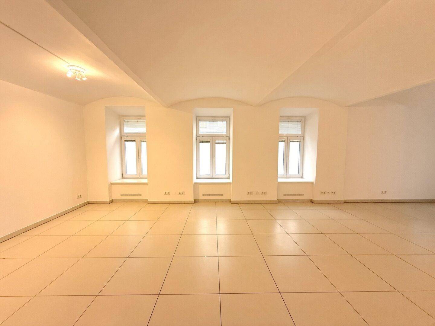 Geräumiger, leerer Raum mit hellen Fliesen, drei Fenstern und gewölbter Decke.