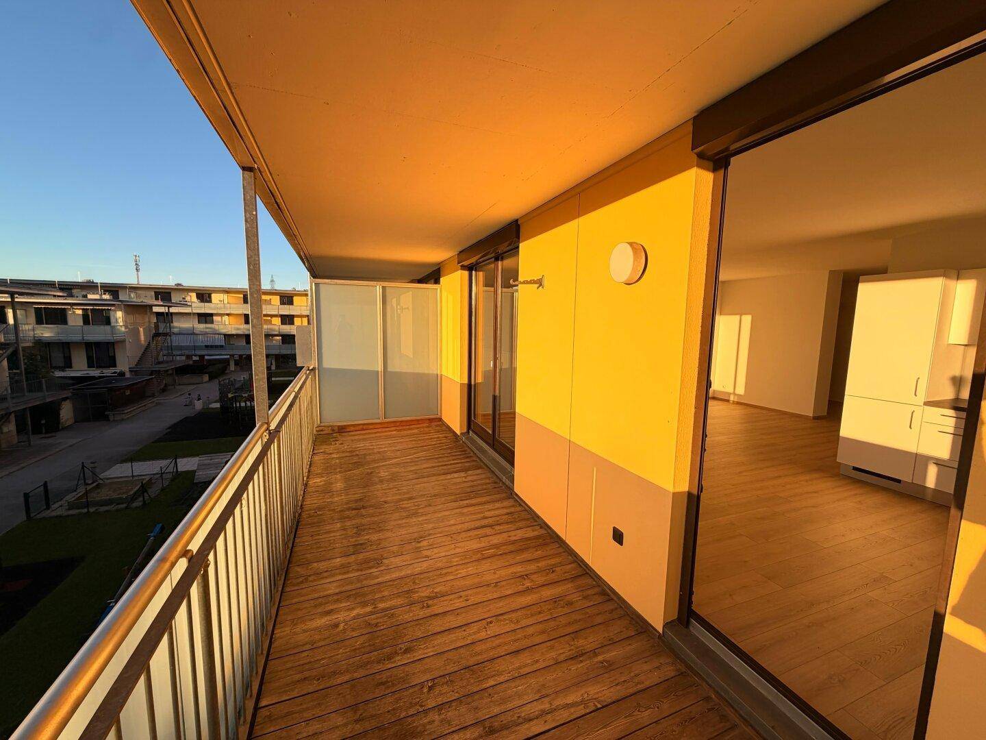 Sonniger Balkon mit Holzboden und Zugang zum Innenbereich, beleuchtet von der Abendsonne.