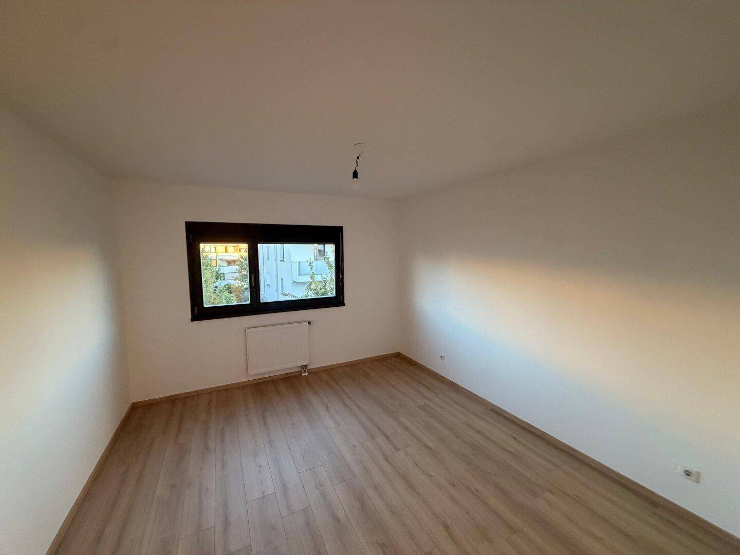 Helles Schlafzimmer mit Laminatboden und großem Fenster, das viel Tageslicht hereinlässt.