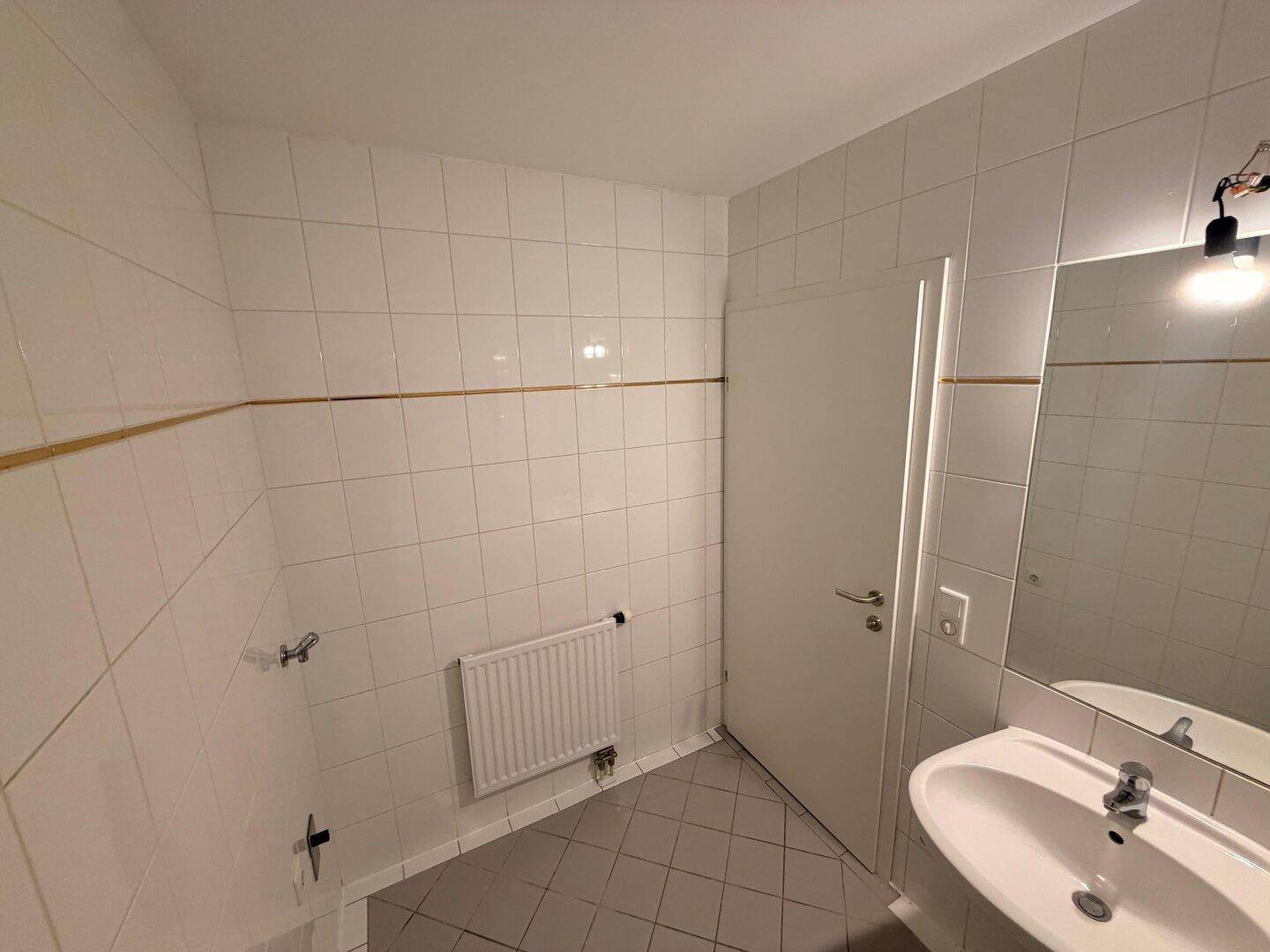 Badezimmer mit weißen Fliesen, Waschbecken, Spiegel und Heizkörper, bereit für die Nutzung.
