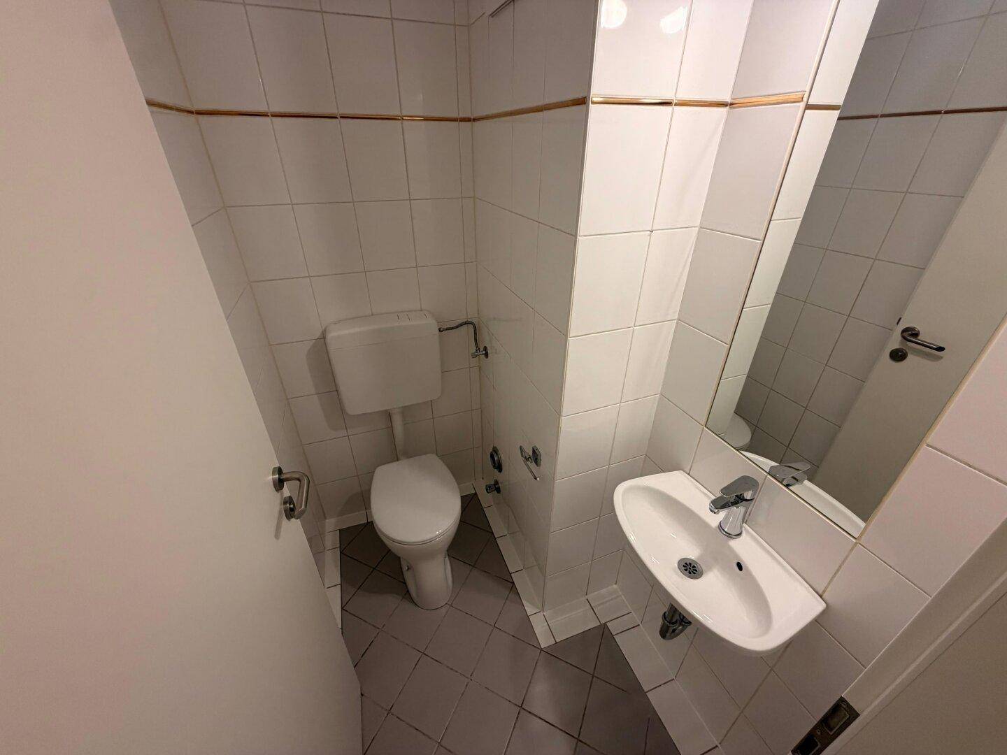 Funktionelles Gäste-WC mit weißen Fliesen, Toilette und kleinem Waschbecken mit Spiegel.