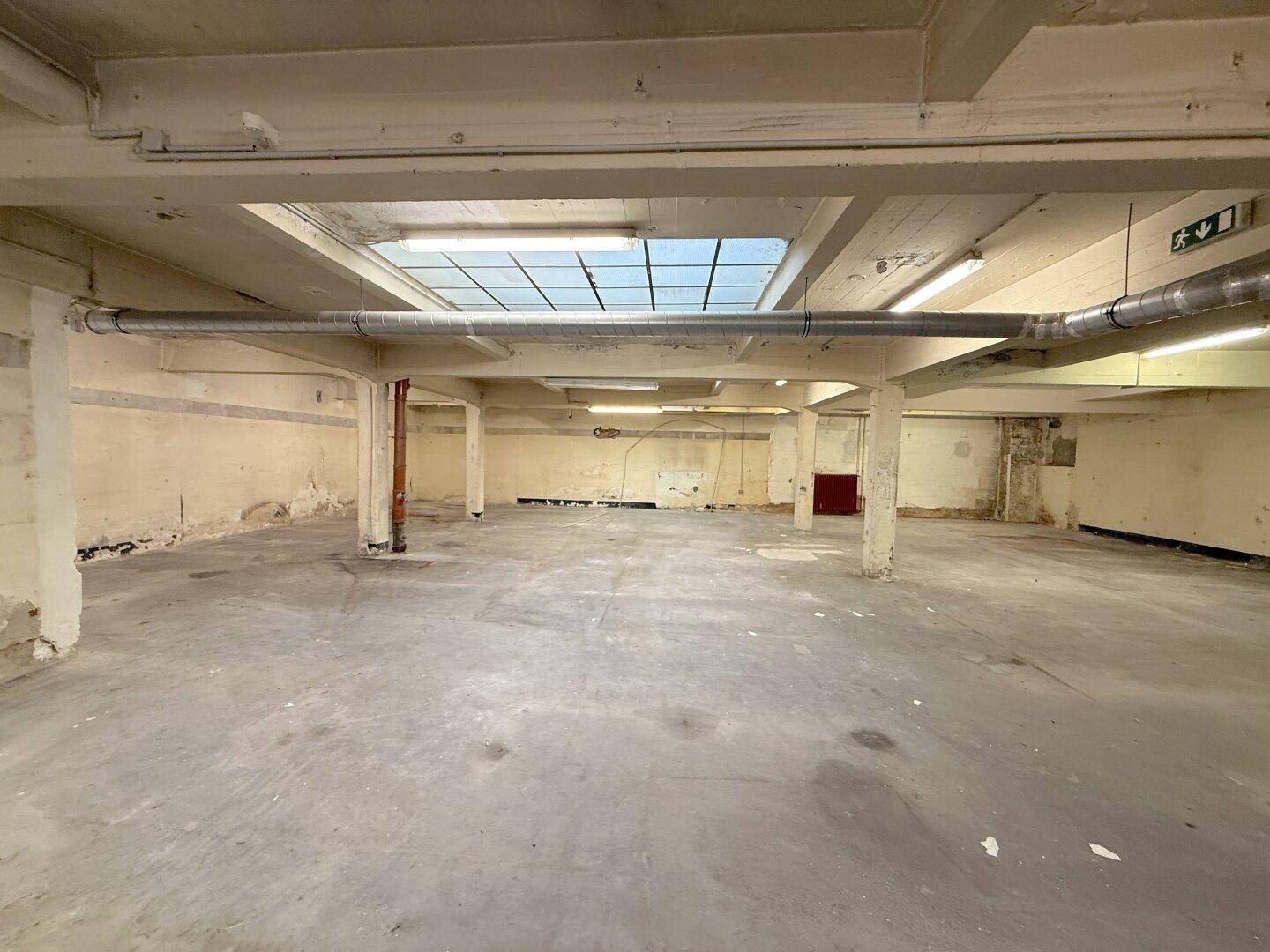 Großer, offener Industrieraum mit Betonboden, freiliegenden Lüftungsrohren und einem großen Oberlicht für Tageslicht.