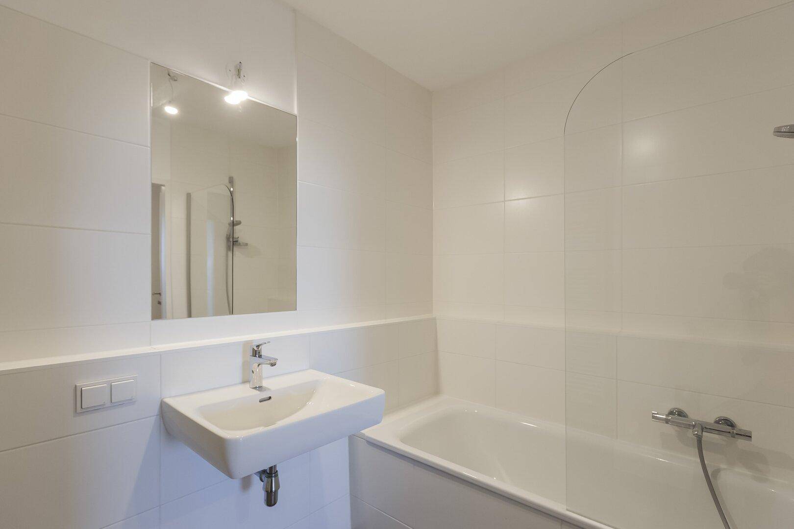 Modernes Badezimmer mit weißen Fliesen, Waschbecken und Badewanne, ausgestattet mit Spiegel.